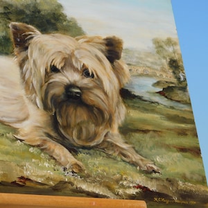 May include: A painting on canvas of a Yorkshire Terrier dog lying down in a field. The dog is brown and white with a long, fluffy coat. The background is a landscape with a bridge and a river.