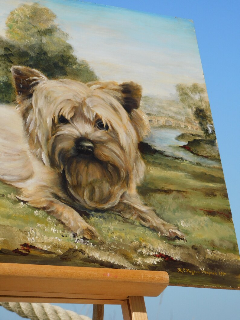 May include: A painting on canvas of a Yorkshire Terrier dog lying down in a field. The dog is brown and white with a long, fluffy coat. The background is a landscape with a bridge and a river.