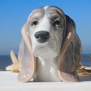 May include: A porcelain figurine of a basset hound dog with brown eyes, a white face, and gray and pink ears. The figurine is set against a blue sky and a white surface.