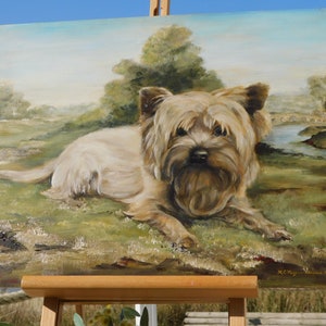 May include: A painting of a small, light brown dog with a fluffy coat, lying down in a grassy field. The dog is looking directly at the viewer. The painting is on an easel and the background is a landscape with a river and a bridge.