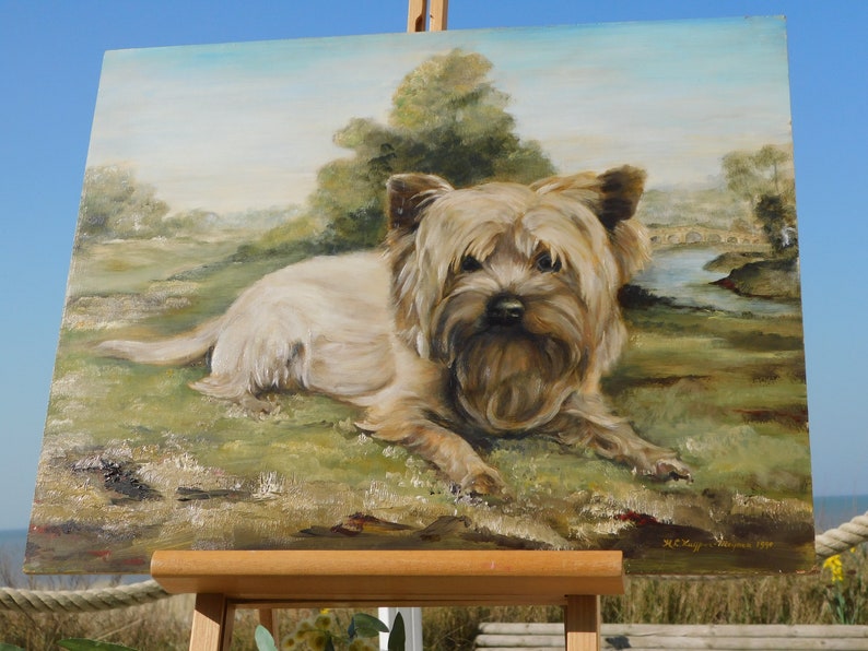 May include: A painting of a small, light brown dog with a fluffy coat, lying down in a grassy field. The dog is looking directly at the viewer. The painting is on an easel and the background is a landscape with a river and a bridge.