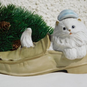 Puede incluir: Una figurita de porcelana blanca de un gato con un sombrero azul, sentado dentro de un zapato marrón.