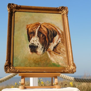 Op de afbeelding: Olieverfschilderij van een Sint-Bernardhond in een gouden, sierlijke lijst. De hond heeft bruine en witte vacht en draagt een halsband. Het schilderij wordt buiten tentoongesteld op een houten ezel met een blauwe lucht op de achtergrond.