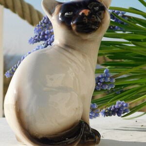 May include: A ceramic figurine of a Siamese cat sitting with its tail curled around its legs. The cat is white with black markings on its face and tail. The figurine is sitting on a white surface with a bouquet of blue flowers in the background.