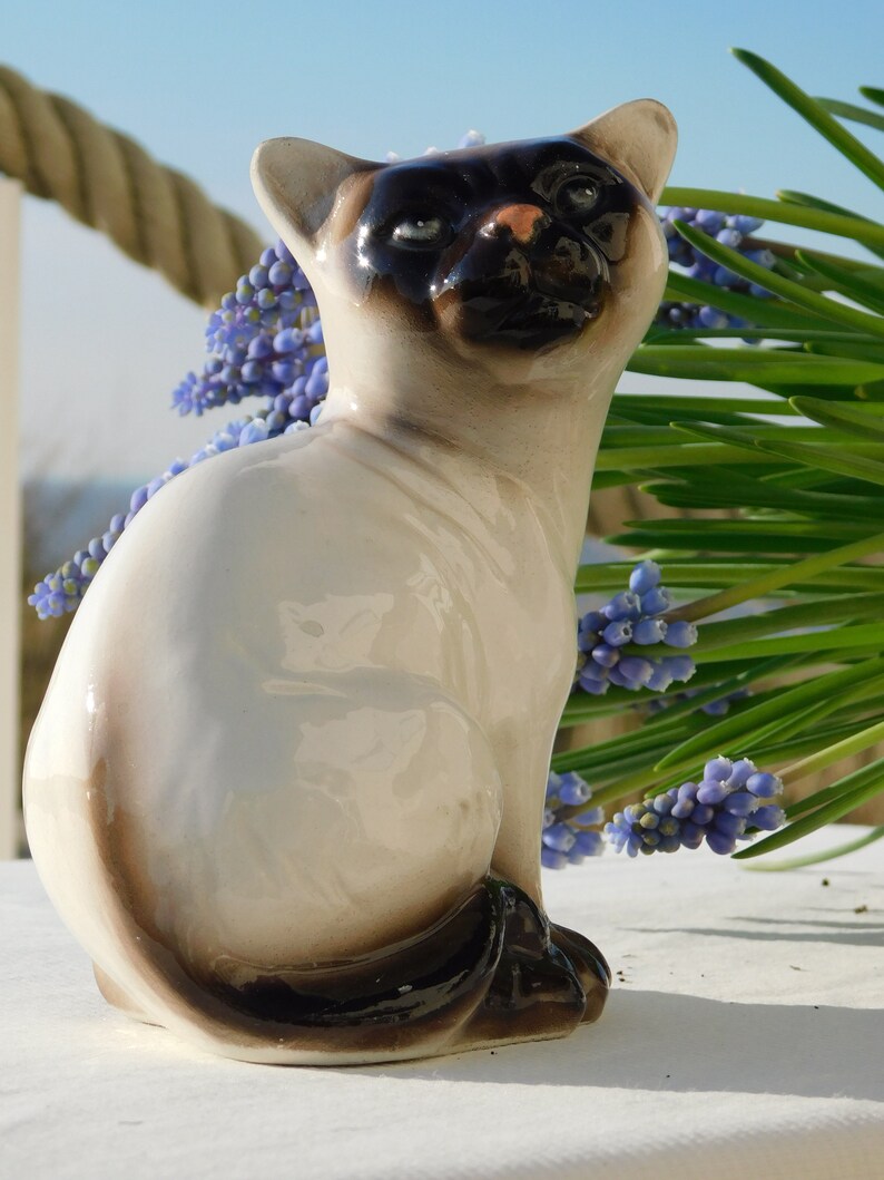 May include: A ceramic figurine of a Siamese cat sitting with its tail curled around its legs. The cat is white with black markings on its face and tail. The figurine is sitting on a white surface with a bouquet of blue flowers in the background.