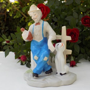 May include: A porcelain figurine of a man wearing a blue overall and a hat, holding a bottle and sitting on a fence with a white dog beside him.