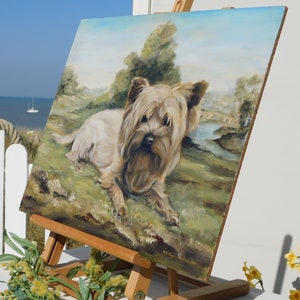 May include: A painting on an easel of a Yorkshire Terrier dog lying in a grassy field with a river and bridge in the background. The painting is framed in a wooden frame and has a light blue sky.