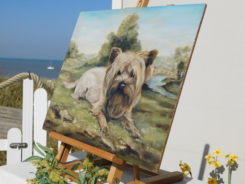 May include: A painting on an easel of a Yorkshire Terrier dog lying in a grassy field with a river and bridge in the background. The painting is framed in a wooden frame and has a light blue sky.
