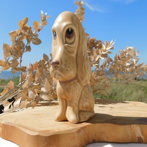 May include: A tan ceramic dog figurine with large, droopy ears and a long snout. The dog is sitting on a wooden surface, with dried foliage in the background. The figurine has brown eyes and a detailed, textured design.