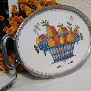 May include: Oval serving tray with silver-toned metal rim and handles. The tray features a white ceramic insert with a hand-painted design of a blue basket filled with pears, apples, and grapes. Two butterflies are also painted on the tray.
