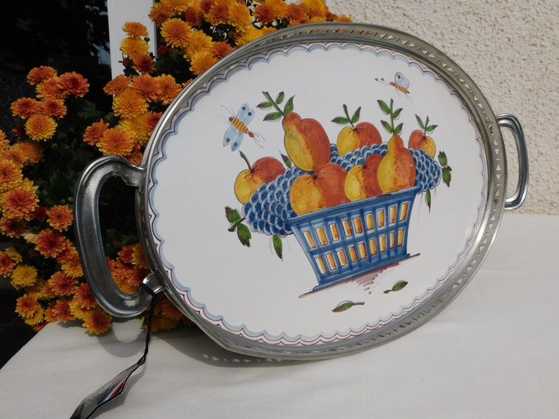 May include: Oval serving tray with silver-toned metal rim and handles. The tray features a white ceramic insert with a hand-painted design of a blue basket filled with pears, apples, and grapes. Two butterflies are also painted on the tray.