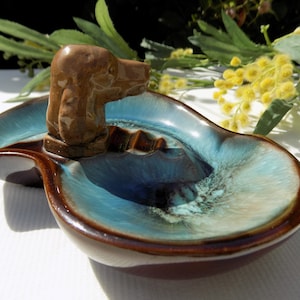 Puede incluir: Un cenicero de cerámica marrón con un esmalte azul y una figurita de perro marrón en la parte superior. El cenicero tiene forma de hoja y tiene una pequeña hendidura para cigarrillos.
