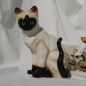 May include: A ceramic figurine of a Siamese cat sitting with its tail curled around its body. The cat is white with black markings and has blue eyes.