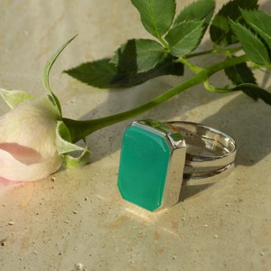 Op de afbeelding: Een zilveren ring met een grote, rechthoekige, smaragdgroene steen. De ring ligt naast een lichtroze roos en groene bladeren. De steen van de ring is ongeveer 2,5 cm lang en 2 cm breed.