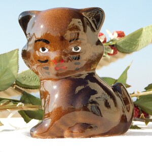Könnte beinhalten: Eine braune Keramik-Katzenfigur mit bemalten schwarzen Augen, einem roten Mund und einem schwarzen Schnurrbart. Die Katze sitzt auf einer weißen Oberfläche mit grünen Blättern und roten Beeren im Hintergrund.