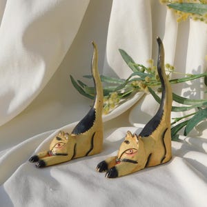 May include: Two hand-carved wooden cat figurines in a stretching pose. The cats are painted yellow with black markings, red eyes, and black paws. The background is a white fabric with greenery.