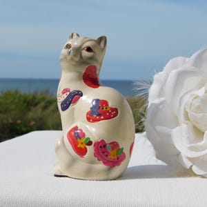 Op de afbeelding: Een crèmekleurig keramieken kattenbeeldje met kleurrijke bloemen- en hoedenontwerpen. De kat zit rechtop met het hoofd iets gedraaid. Rechts van de kat staat een grote witte roos.
