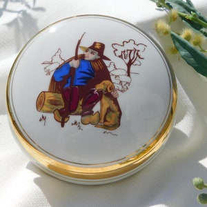 Könnte beinhalten: Eine weiße Porzellan-Schmuckdose mit goldfarbenem Rand. Der Deckel zeigt eine handgemalte Szene eines Mannes in einem blauen Hemd und braunen Hosen, der auf einem Baumstamm mit einem Hund sitzt. Die Szene spielt in einem Wald mit Bäumen und Büschen.