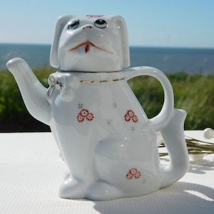 Op de afbeelding: Witte porseleinen theepot in de vorm van een hond met rode bloemmotieven. De theepot heeft een deksel in de vorm van een hondenkop.