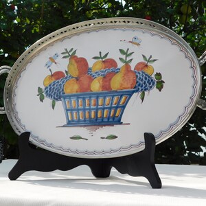 May include: Oval serving tray with silver-toned rim and handles. The tray features a hand-painted design of a blue basket filled with yellow, orange, and red fruit. The fruit is surrounded by green leaves and blue grapes.
