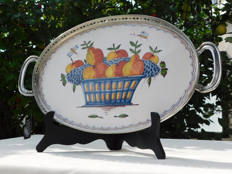 May include: Oval serving tray with silver-toned rim and handles. The tray features a hand-painted design of a blue basket filled with yellow, orange, and red fruit. The fruit is surrounded by green leaves and blue grapes.