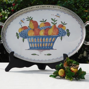 May include: An oval silver serving tray with a white background and a hand-painted design of a blue basket filled with pears and apples. The tray has two silver handles and a decorative silver rim.