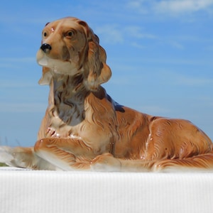 Op de afbeelding: Een keramiek beeldje van een bruine hond die ligt. De hond heeft een lange snuit en hangende oren. Het beeldje is gedetailleerd met vacht en heeft een glanzende afwerking.