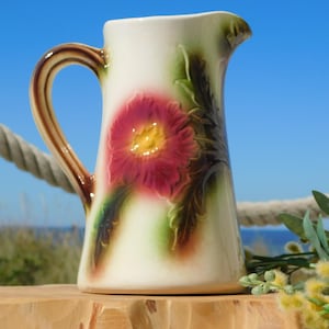 Puede incluir: Una jarra de cerámica blanca con asa marrón y un diseño floral. La jarra presenta una gran flor rosa con un centro amarillo y hojas verdes.