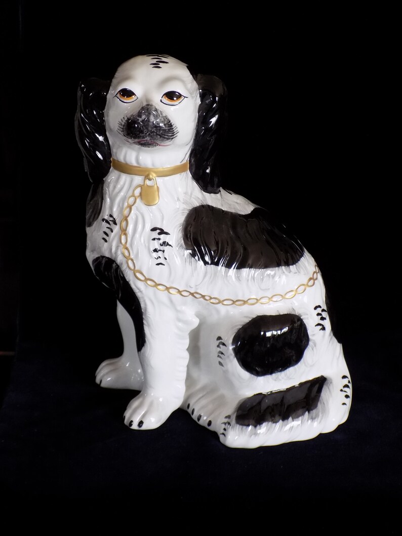 K&ouml;nnte beinhalten: Eine schwarz-wei&szlig;e Porzellanfigur eines Hundes mit einem goldenen Kettenhalsband. Der Hund sitzt mit dem Kopf zur Seite geneigt und die Ohren h&auml;ngen herunter.