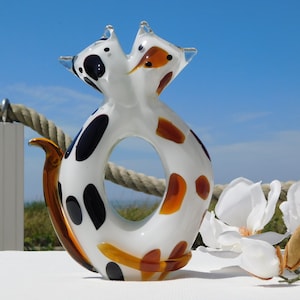 Puede incluir: Una escultura de vidrio blanco de dos gatos entrelazados, con manchas negras y marrones. La escultura está sobre una superficie blanca con una cuerda y flores en el fondo.