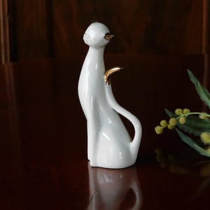 Op de afbeelding: Een witte keramische kattenfiguur met gouden accenten op de oren en staart. De kat zit met een lange, gebogen staart. De figuur staat op een donkerbruin oppervlak.