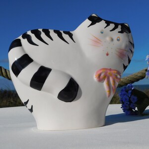 Op de afbeelding: Een witte keramische kattenfiguur met zwarte strepen en een gestreepte staart. De kat heeft roze wangen, blauwe ogen en een roze strik. De figuur staat op een wit oppervlak met blauwe bloemen op de achtergrond.