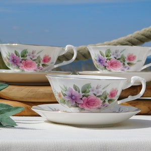 May include: Three white teacups and saucers with a floral pattern featuring pink, purple, and white flowers. The cups and saucers have a gold trim.