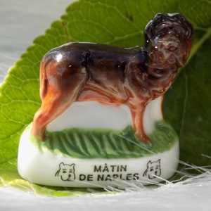 May include: A brown and white porcelain figurine of a Neapolitan Mastiff dog. The figurine is standing on a green base with the words "MâtIN DE NAPLES" written on it.