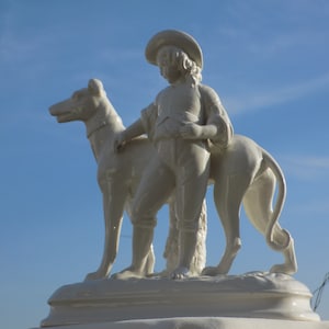 Könnte beinhalten: Weiße Porzellanfigur eines jungen Menschen in einem Hut, der einen Windhund hält.