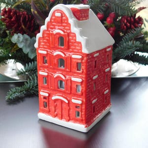 May include: A red ceramic house-shaped candle holder with a white roof and trim. The house has multiple windows and a chimney. The background features evergreen branches and red berries, suggesting a festive theme.