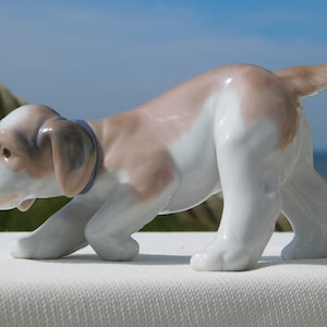 Op de afbeelding: Een porseleinen beeldje van een bruine en witte puppy met een blauwe halsband, die met zijn kop naar beneden loopt. Het beeldje staat op een wit oppervlak.
