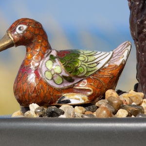 Puede incluir: Una pequeña figurita de pato de cloisonné, finamente detallada, con un diseño de esmalte marrón, rojo, verde y blanco. El pato está sentado en un lecho de pequeñas piedras grises.