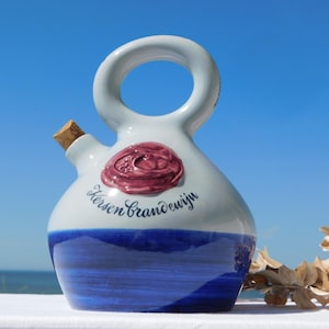 Op de afbeelding: Een witte keramische kan met een rond lichaam en een cirkelvormig handvat aan de bovenkant. De basis is beschilderd met een donkerblauwe horizontale band. Een verhoogd, donkerroze kersvormig embleem staat boven de zwarte tekst Kersen Brandewijn.