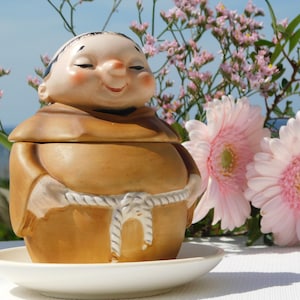 Op de afbeelding: Een keramiek beeldje van een glimlachende monnik die een bruine robe draagt met een touwgordel. Het beeldje houdt een container met deksel vast. Het beeldje staat op een witte schotel. De achtergrond is een wazig beeld van roze bloemen.
