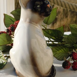 May include: A white porcelain figurine of a Siamese cat sitting with its tail curled around its legs. The cat has black markings on its face and tail.