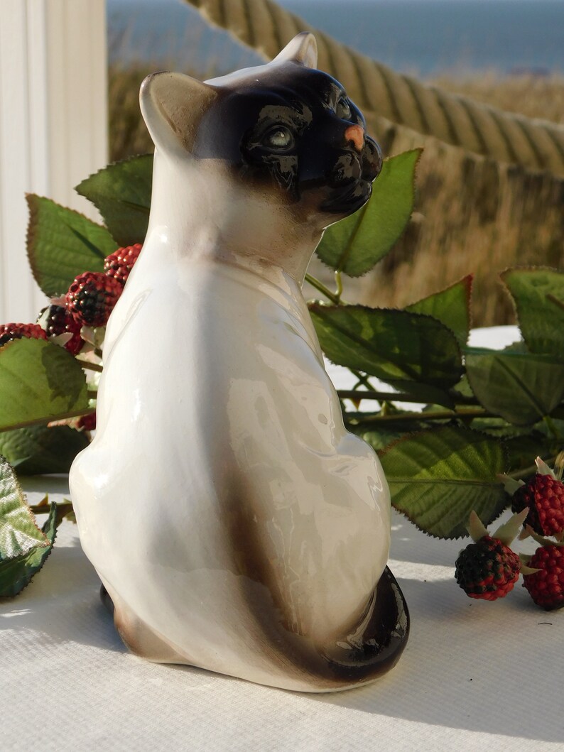 May include: A white porcelain figurine of a Siamese cat sitting with its tail curled around its legs. The cat has black markings on its face and tail.