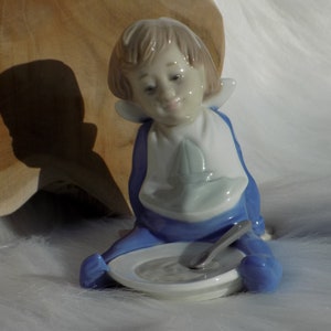 Op de afbeelding: Een porseleinen beeldje van een kind dat zit met een kom en een lepel. Het kind draagt een blauwe en witte outfit met een slabbetje. Het beeldje zit in een zittende positie met een kom en een lepel voor zich.