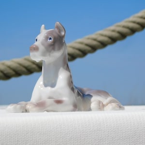 May include: A glossy porcelain figurine of a dog in a lying position. The dog is primarily white with soft grey patches on its head, ears, and back. It has pointed ears, a smooth finish, and a focused expression with small, dark, painted eyes.