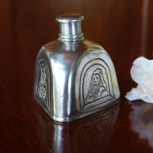 Peut inclure: Flacon de parfum argenté de forme rectangulaire arrondie. La bouteille présente des images gravées d'un homme barbu coiffé d'un couvre-chef. Il possède un petit bouchon cylindrique. Fond en bois sombre.