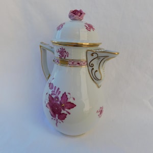Op de afbeelding: Een witte porseleinen koffiepot met magenta bloemmotieven, gouden randaccenten en een deksel bekroond met een gebeeldhouwde roze roos. De kan is voorzien van een gebogen handvat en een decoratieve tuit met gedetailleerde botanische patronen.