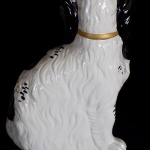 K&ouml;nnte beinhalten: Eine wei&szlig;e und schwarze Keramikfigur eines sitzenden Hundes, der den Kopf zur Seite dreht. Der Hund tr&auml;gt ein goldenes Halsband und hat schwarze Markierungen am K&ouml;rper.