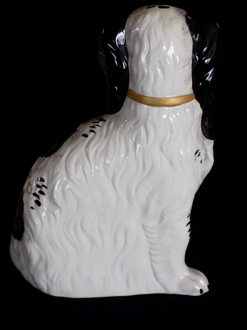 K&ouml;nnte beinhalten: Eine wei&szlig;e und schwarze Keramikfigur eines sitzenden Hundes, der den Kopf zur Seite dreht. Der Hund tr&auml;gt ein goldenes Halsband und hat schwarze Markierungen am K&ouml;rper.