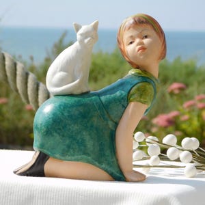 Könnte beinhalten: Eine Porzellanfigur eines Mädchens in einem grünen Kleid mit einer weißen Katze, die auf ihrem Rücken sitzt. Das Mädchen kniet und schaut zur Seite. Die Figur steht auf einer weißen Oberfläche.