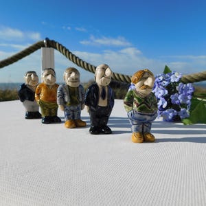 Op de afbeelding: Een rij van vijf keramische beeldjes van mannen, elk met een unieke outfit en uitdrukking. De figuren staan op een wit oppervlak met een touw en blauwe bloemen op de achtergrond. De lucht is blauw met een paar wolken.